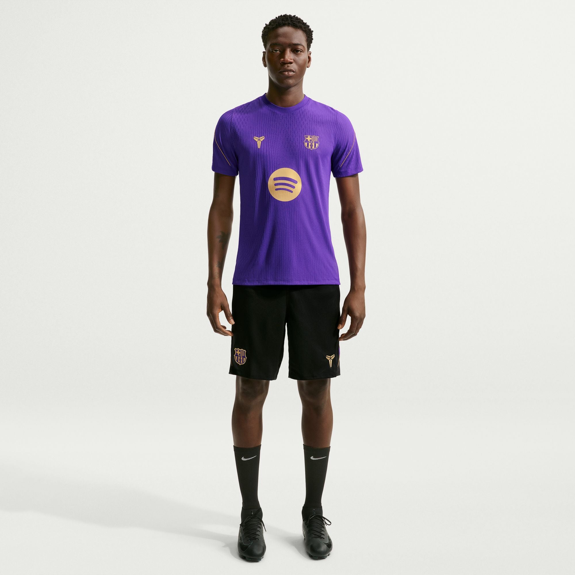 A male model stands against a light gray studio background wearing a purple short-sleeve soccer jersey with gold logos, black shorts, black socks, and black cleats. The jersey has a large circular gold logo on the front and a smaller crest on the chest.
