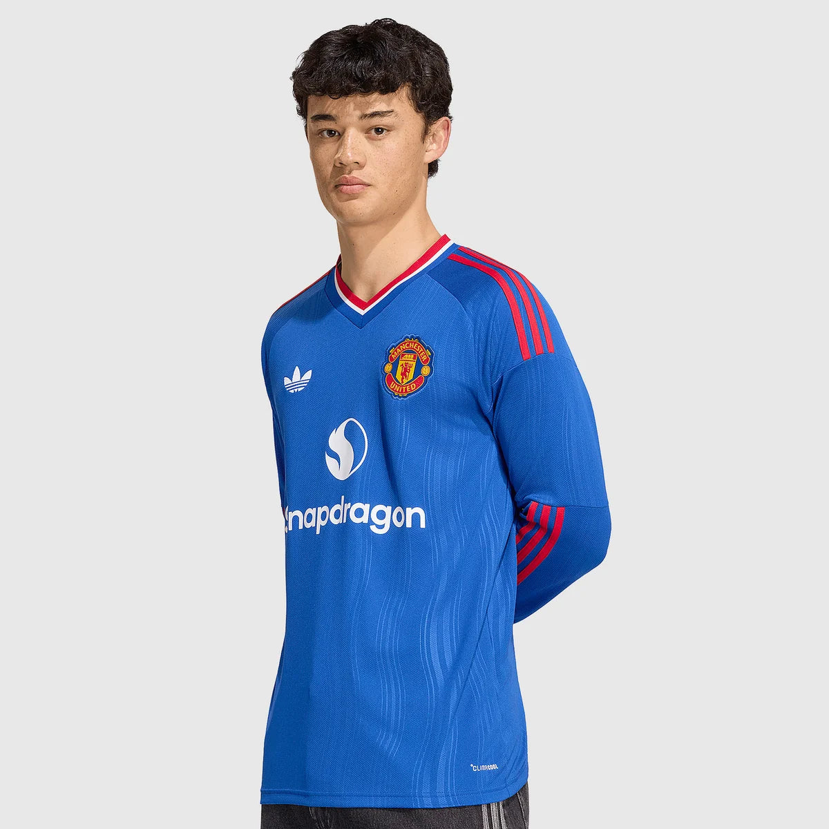 A male model wearing a blue Manchester United Adidas soccer jersey with red stripes and white sponsor logos, set against a plain light gray background.
