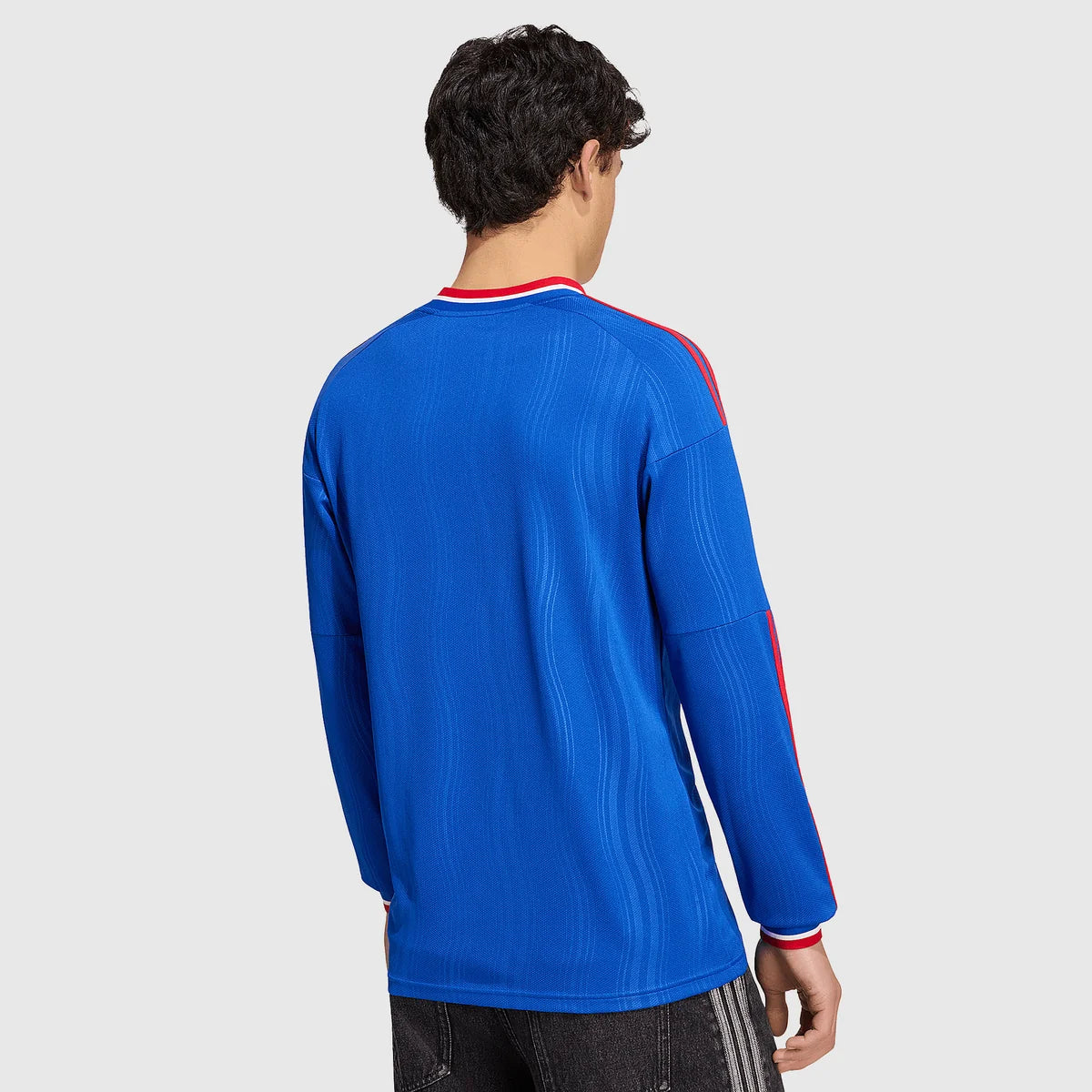 A man viewed from the back wearing a royal blue long-sleeve athletic jersey with red and white trim on the collar and sleeve, paired with dark gray pants, against a light gray studio background.