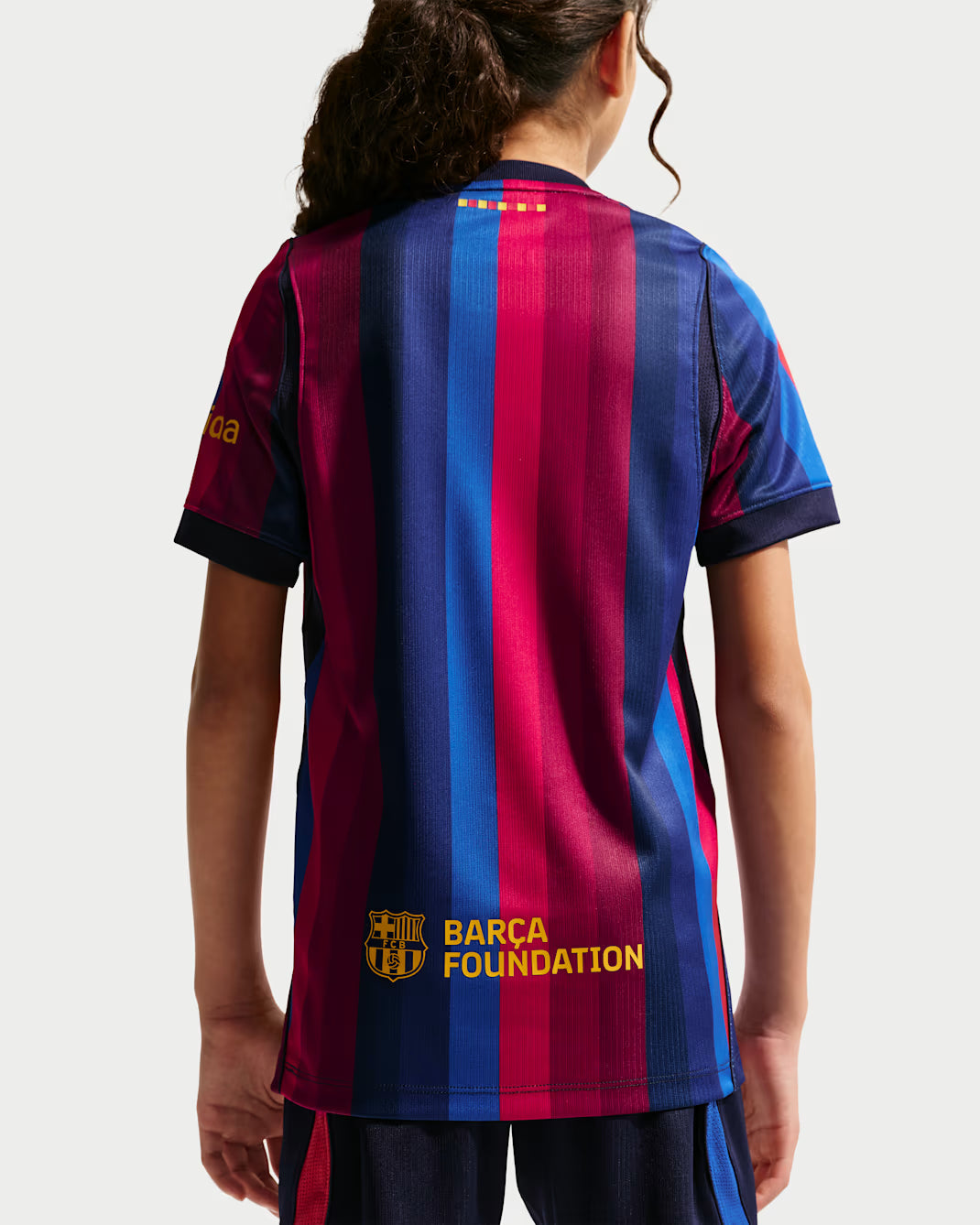 A child wearing a FC Barcelona home soccer jersey, shown from the back. The jersey features the classic vertical stripes in navy blue, red, and blue, with gold accents including the FC Barcelona crest, 'BARCA FOUNDATION' text, and team stars at the collar.