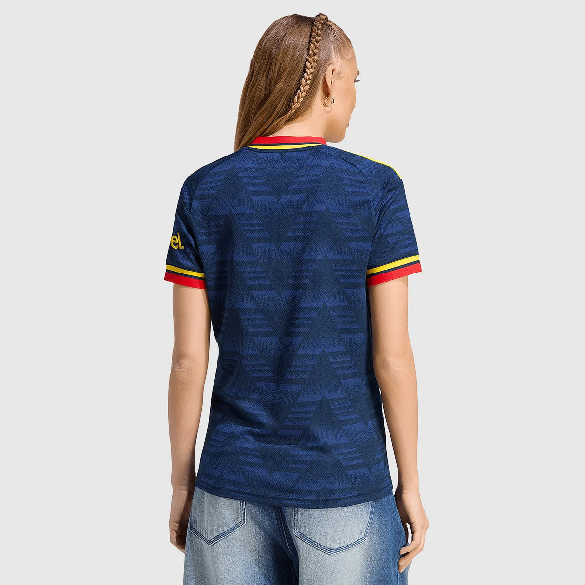 A woman viewed from the back wearing a dark blue patterned short-sleeve soccer jersey with red and yellow trim on the collar and sleeve cuffs, paired with light blue jeans, against a light gray studio background.