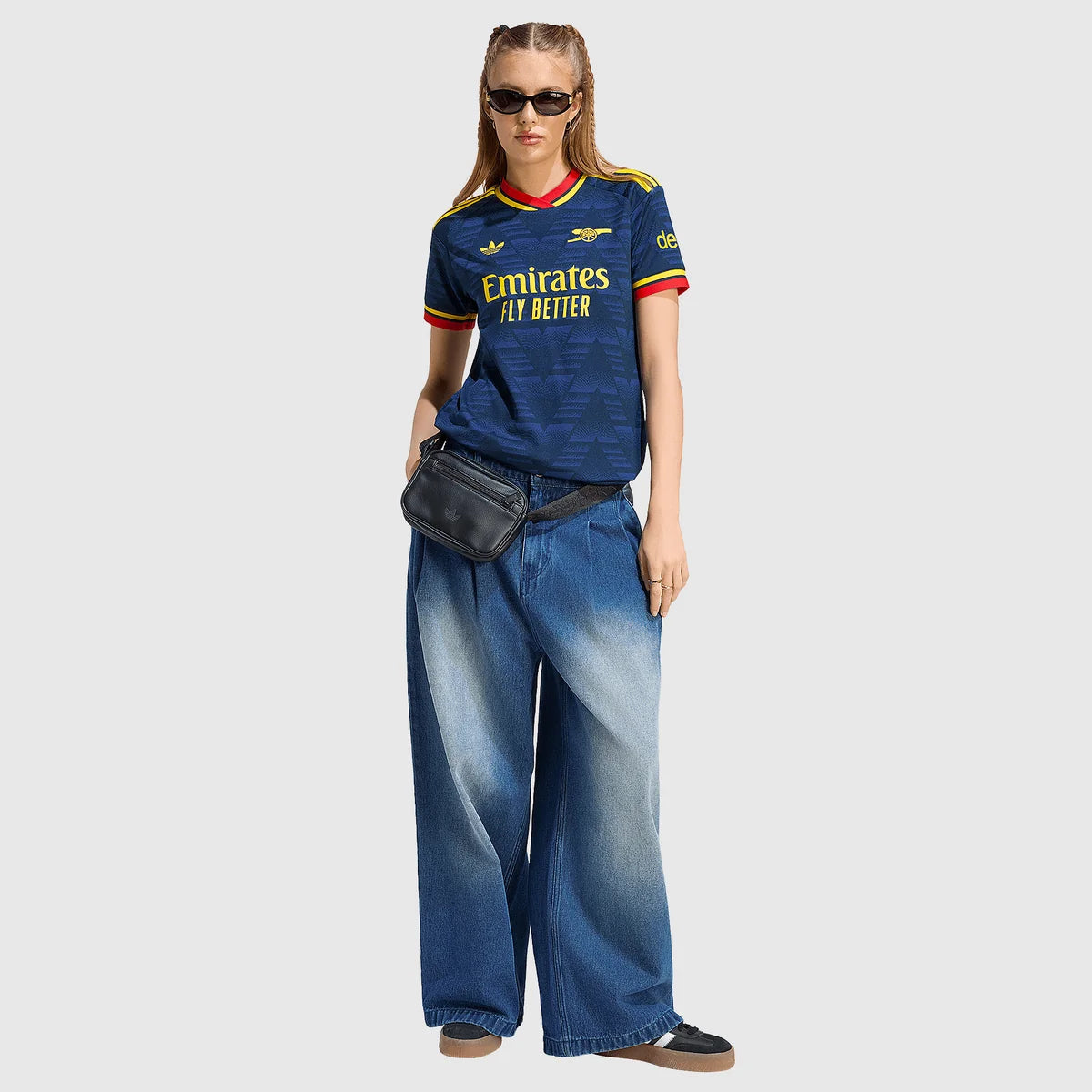 A woman modeling a navy blue Arsenal football shirt with yellow and red trim, paired with wide-leg faded blue jeans, a black belt, and a black fanny pack, against a light gray background.