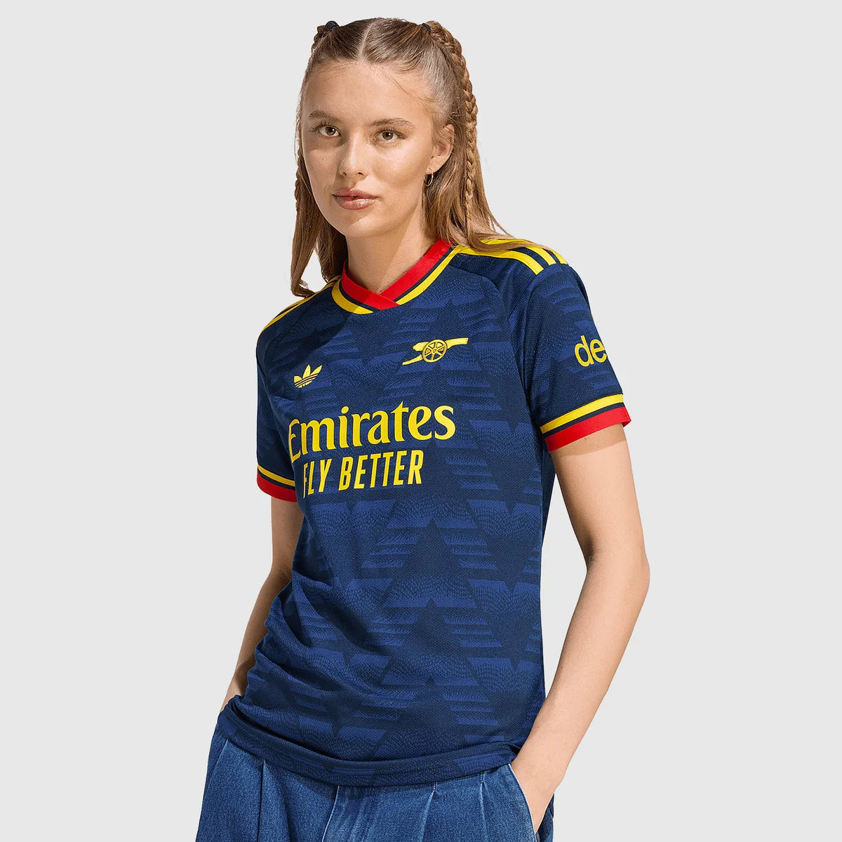 A woman modeling a navy blue Arsenal football jersey with yellow and red trim, featuring the Emirates Fly Better sponsor logo on the chest, set against a solid light gray background.