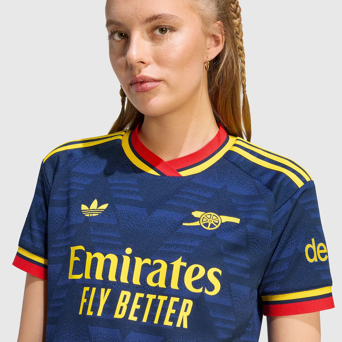 A close-up of a woman wearing a navy blue Arsenal football jersey with yellow and red accents, featuring the Adidas logo, Arsenal crest, and Emirates Fly Better sponsor text.