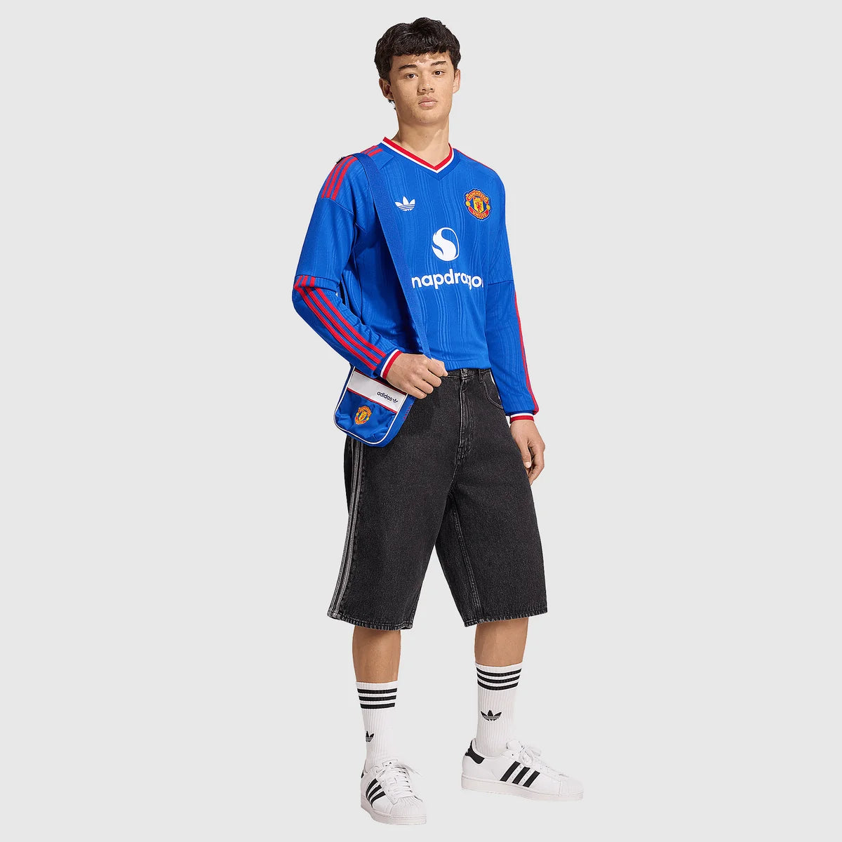 A model wearing a blue Manchester United long-sleeve soccer jersey by Adidas, styled with dark denim shorts, white socks, and white Adidas Superstars on a light gray background.